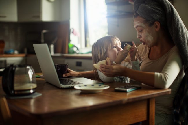 Busy mom with hair wrapped in towel wearing a skin care face mask while working at her laptop with daughter on her lap | Vitacost.com/Blog