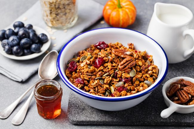 Homemade cardamom maple granola in a bowl surrounded by pecans, syrup and pumpkin | Vitacost.com/Blog