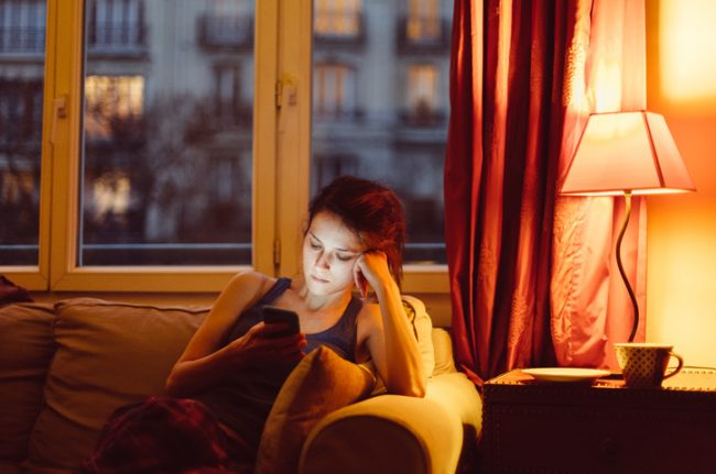 Woman Suffering From Sedentary Lifestyle Sitting on Couch Staring Sadly at Phone | Vitacost Blog 