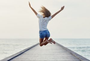 Woman Following Tips to Increase Energy Jumping With Arms Up on Dock | Vitacost.com/blog