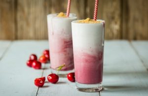 2 Cherry Cheesecake Smoothies in Tall Glasses with Straws & Cherries on White Wood Table #recipe | Vitacost.com/blog