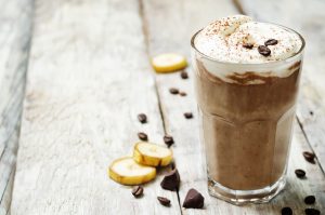 Espresso Protein Smoothie in Glass with Froth, Coffee Beans & Banana Slices on Wood Table | Vitacost.com/blog