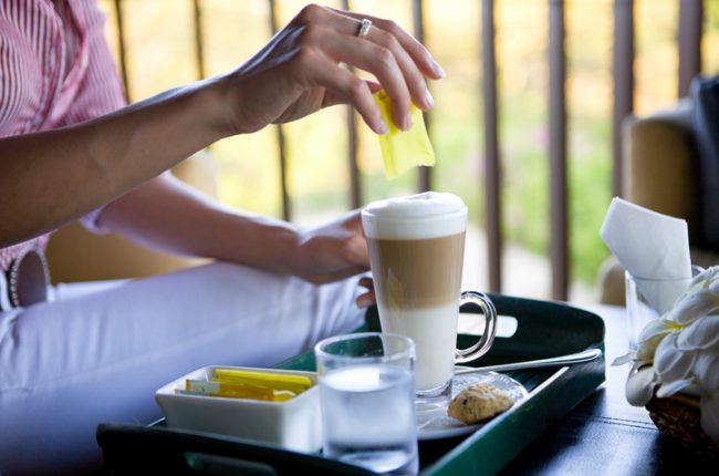 Woman at Coffee Shop Pouring Artificial Sweeteners Into a Latte | Vitacost.com/blog