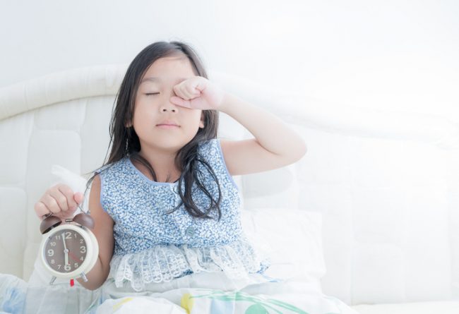 How Much Sleep Do Kids Really Need? Groggy Young Girl Rubbing Her Eyes in Bed Due to Lack of Sleep | Vitacost.com/blog