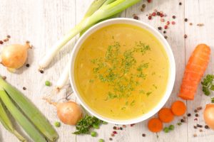 White Bowl of Turkey Carcass Soup Stock on White Wood with Carrots, Onions & Leaks Around Bowl | Vitacost.com/blog
