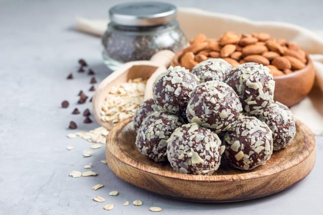 Healthy Chocolate Date Truffles  Wood Bowl of Healthy Chocolate Date Truffles with Toasted Coconut Coating & Almonds, Oats in Background #recipe | Vitacost.com/blog