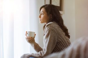 Woman Holding Cup of Tea & Looking Out Window Needs These 2 Herbs for Winter Mood Support – Learn More | Vitacost.com/blog