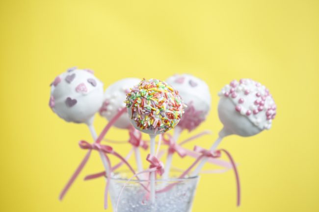 Cheesecake Pops for Galentine’s Day 5 White Chocolate Galentine’s Day Cheesecake Pops with Sprinkles in Clear Cup with Yellow Background #recipe | Vitacost.com/blog