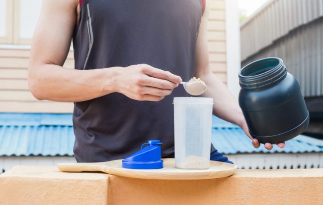Athletic Man Reaping Whey Protein Benefits by Scooping Powder into Smoothie Shaker | Vitacost.com/blog