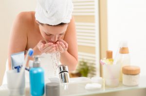 Towel-Clad Woman at Bathroom Sink Rinsing Face After Learning How to Exfoliate | Vitacost.com/blog