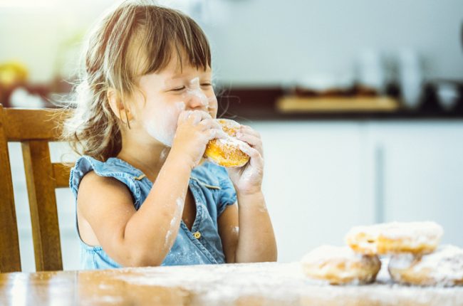 Little Girl Enjoying One of the Unhealthiest Foods for Kids Biting into Powdered Sugar-Covered Donut | Vitacost.com/blog