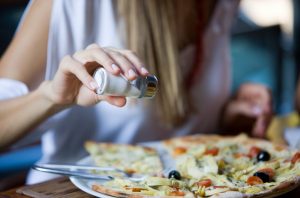 Woman Unconcerned About Blood Pressure Health Sprinkling Extra Salt on Pizza | Vitacost.com/blog