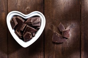 White Heart-Shaped Bowl Filled with Squares & Chunks of Dark Chocolate on Wooden Table |