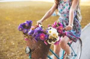 Woman Riding Bicycle With Colorful Flowers in Front Basket Practicing Healthy Living Tips for Summer | Vitacost.com/blog