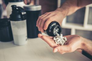 A Man’s Hands Pour Out a Bottle of the Best Vitamins for Men With Protein Shaker Cup in Background | Vitacost.com/blog