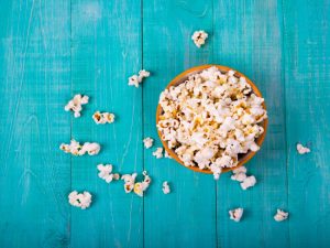 Bowl of Popcorn Surrounded by Scattered Kernels on Teal Wooden Table Representing Gluten Free Snack Ideas | Vitacost.com/blog