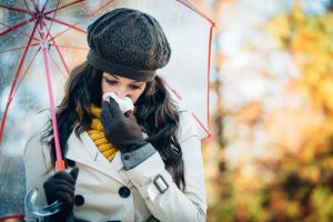 Woman in Coat with Umbrella Blowing Nose into Tissue as Autumn Season Change Triggered her Allergies | Vitacost.com/blog