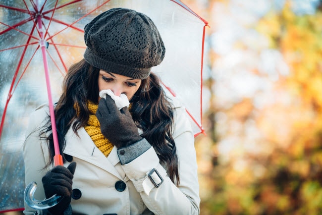 Woman in Coat with Umbrella Blowing Nose into Tissue as Autumn Season Change Triggered her Allergies | Vitacost.com/blog