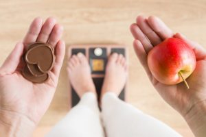 How to Break a Weight Loss Plateau Represented by Person Standing on Scale Holding Apple in One Hand and Candy in Other | Vitacost.com/blog