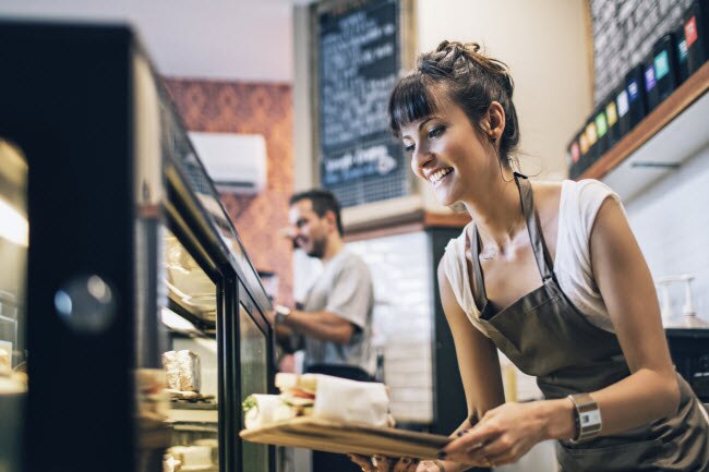Women-Owned Business Leaders in Natural Foods  Smiling Woman Working Behind Counter of Women Owned Business Bakery and Deli | Vitacost.com/blog