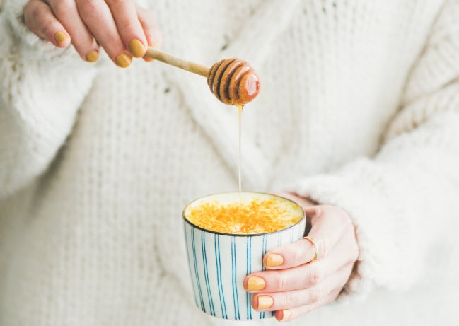 10 Must-Have Natural Products for Healthy Living Woman in White Sweater Practicing Healthy Living Pouring Raw Honey Into Mug of Tea | Vitacost.com/blog
