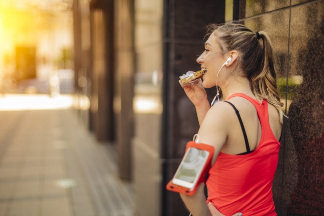 Athletic Woman in Red Workout Top Standing Against City Building Eating a Snack Bar | Vitacost.com/blog