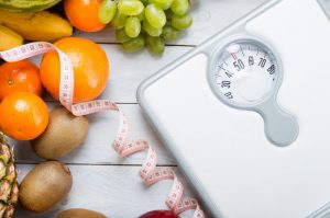 White Scale, Coiled Measuring Tape and Fruit on White Wooden Table to Represent Tips for Keto Not Working | Vitacost.com/blog