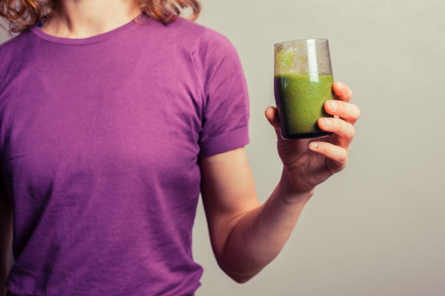 Torso View of Woman in Purple T-Shirt Who Knows What's Good for a Hangover Holding Glass of Green Juice | Vitacost.com/blog