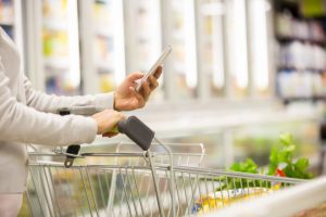 Woman Following Budget Tips Learning How to Eat Healthy Pushing Grocery Cart While Calculating Food Cost on Phone Calculator | Vitacost.com/blog