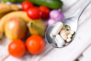 Close-up Image of Vitamins and Supplements on a Spoon with Faded Fruits and Vegetables in Background | Vitacost.com/blog