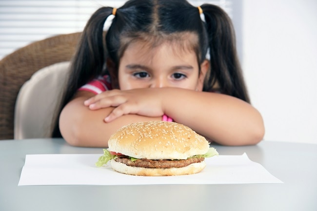 How Much Protein Do Kids Needs + Why it’s Important  Girl Sitting at Table Staring Hamburger to Show How Much Protein Kids Need | Vitacost.com/blog