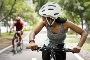 Helmet-Clad Woman Riding a Bike Celebrating National Bike Month Looking Back at Partner Riding Behind Her | Vitacost.com/blog