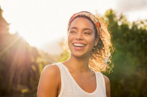 Smiling Woman Who Sweat Proofed Her Summer Beauty Routine Standing in Woods with Sun Shining Behind Her | Vitacost.com/blog