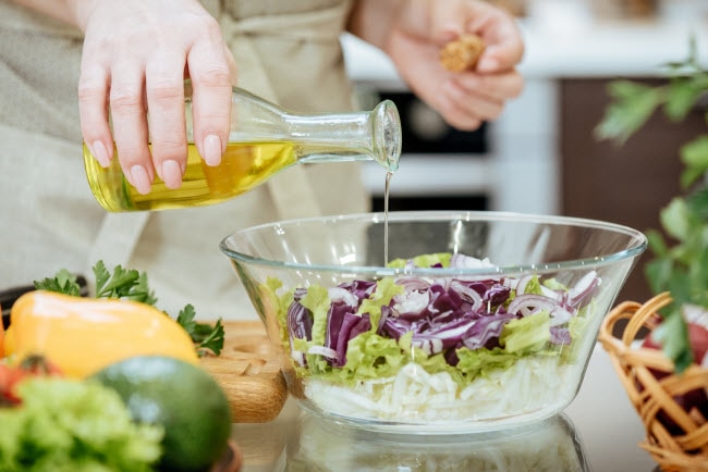 Woman Pouring Oil Over Salad in Glass Bowl as Example of One of Many MCT Oil Uses | Vitacost.com/blog