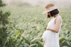 Pregnant Woman Who Followed Holisitic Fertility Tips Wearing White Dress and Sunhat Standing in Green Field | Vitacost.com/blog