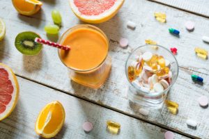 Two Glasses Filled With Essential Vitamins for Summer Health and Juice Surrounded by Fruit and Capsules on Wooden Table | Vitacost.com/blog