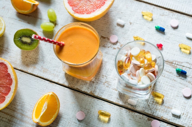 Two Glasses Filled With Essential Vitamins for Summer Health and Juice Surrounded by Fruit and Capsules on Wooden Table | Vitacost.com/blog