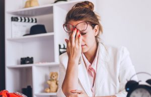 Young Woman With Glasses Sitting at Desk Holding Forehead in Pain as She's Experiencing Women's Health Issues | Vitacost.com/blog