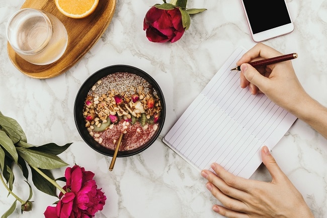 Person with Smoothie Bowl Writing Plant-Based Meal Plan in Notebook | Vitacost.com/Blog