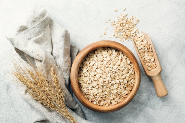 The Benefits of Whole Grains Represented by Wooden Bowl of Oats with Scoop and Napkin on Gray Marble Surface | Vitacost.com/blog