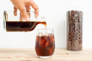 How to Make Cold Brew Coffee Represented by Jar of Cold Brew Pouring into Glass of Ice on Wooden Table Beside Tall Canister of Coffee Beans | Vitacost.om/blog