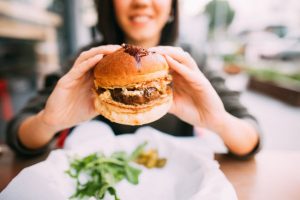 Question of 'Is Vegan Meat Healthy' Represented by Zoomed in Shot of Veggie Burger Held by Slightly Faded Out Smiling Woman at Cafe | Vitacost.com/blog