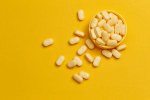 What is B12 and Why is B12 Important Represented by Yellow Vitamin Tablets Overflowing from Bottle Cap on Yellow Surface | Vitacost.com/blog