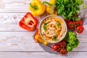 Overhead View of Bowl of Freshly Made Hummus with Red and Yellow Peppers and Tomatoes on Wood Surface for Low-Sugar Snack Ideas | Vitacost.com/blog