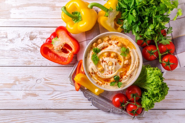 Overhead View of Bowl of Freshly Made Hummus with Red and Yellow Peppers and Tomatoes on Wood Surface for Low-Sugar Snack Ideas | Vitacost.com/blog