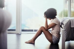 Woman Experiencing Signs of Stress Sitting on Floor at End of Bed Holding Her Forehead in Pain | Vitacost.com/blog