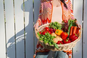 Torso View of Woman Following Plant-Based Nutrition Tips Holding Basket of Fresh Produce in Front of White Picket Fence | Vitacost.com/blog