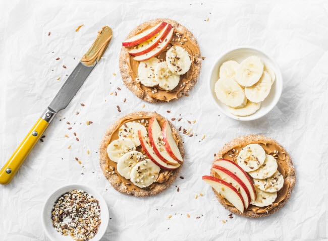 Sliced Bananas and Apples Rice Cakes with Knife on White Marble Surface to Represent Best Prebiotic Foods for Gut Health | Vitacost/com/blog