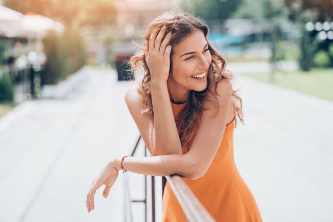 How to Get Glowing Skin Naturally Long-Haired Brunette Woman in Orange Dress Who Learned How to Get Glowing Skin Naturally Laughing While Leaning on Railing in a Park | Vitacost.com/blog