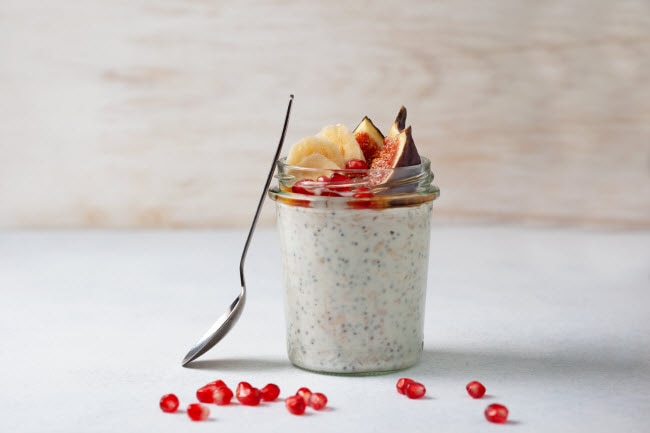 How to Make Overnight Oats Exemplified by Jar of Oatmeal Topped by Figs and Bananas on White Surface With Scattered Pomegranate Arils | Vitacost.com/blog